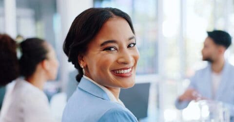 Woman in a light blue blazer smiling at the camera in a bright office setting, with two colleagues blurred in the background.