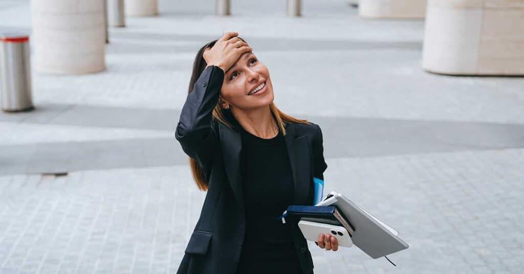 Is This It? What An 18-Year-Old Taught Me About Success Woman in business attire smiles while holding notebooks and a phone, touching her forehead with one hand, standing outdoors on a paved area with pillars in the background.