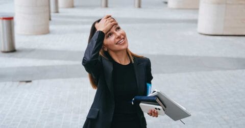 Woman in business attire smiles while holding notebooks and a phone, touching her forehead with one hand, standing outdoors on a paved area with pillars in the background.