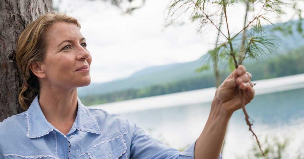 What If You Didn’t Need A New Life? Woman in a blue shirt stands by a tree, holding a pine branch, with a lake and mountains in the background on a cloudy day.
