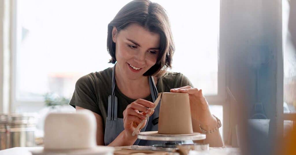 When Was The Last Time You Surprised Yourself? A woman shapes a clay pot on a pottery wheel, using a small tool to smooth the surface. She is wearing an apron and smiling while working.