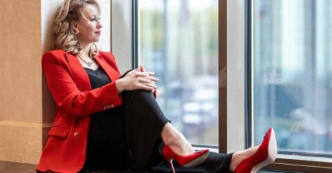 Woman in a red blazer and red high heels sits on the floor by a large window, looking outside with her hands resting on her knee.