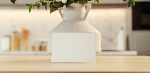 A blank white rectangular card leaning against a speckled ceramic vase on a wooden surface.